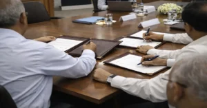 Hands signing official documents during an MoU between NHAI and Konkan Railway in a government office setting.