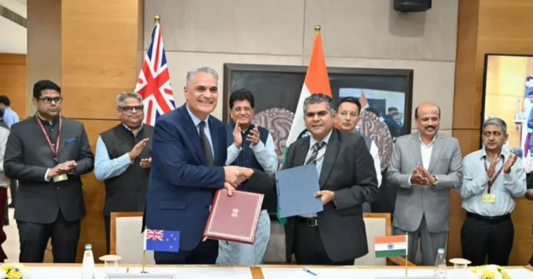 Indian and New Zealand representatives shake hands and exchange documents during a bilateral engagement, with both countries' national flags displayed.