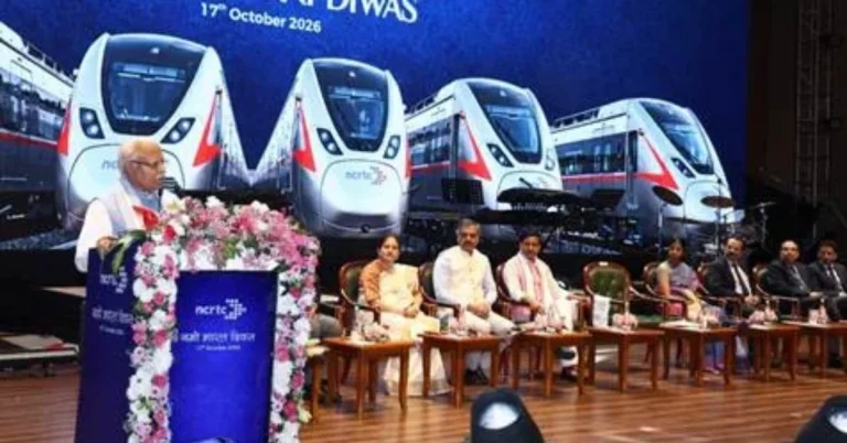 A speaker addresses Namo Bharat Diwas 2026 on stage, with officials seated beside a large backdrop displaying Namo Bharat trains.